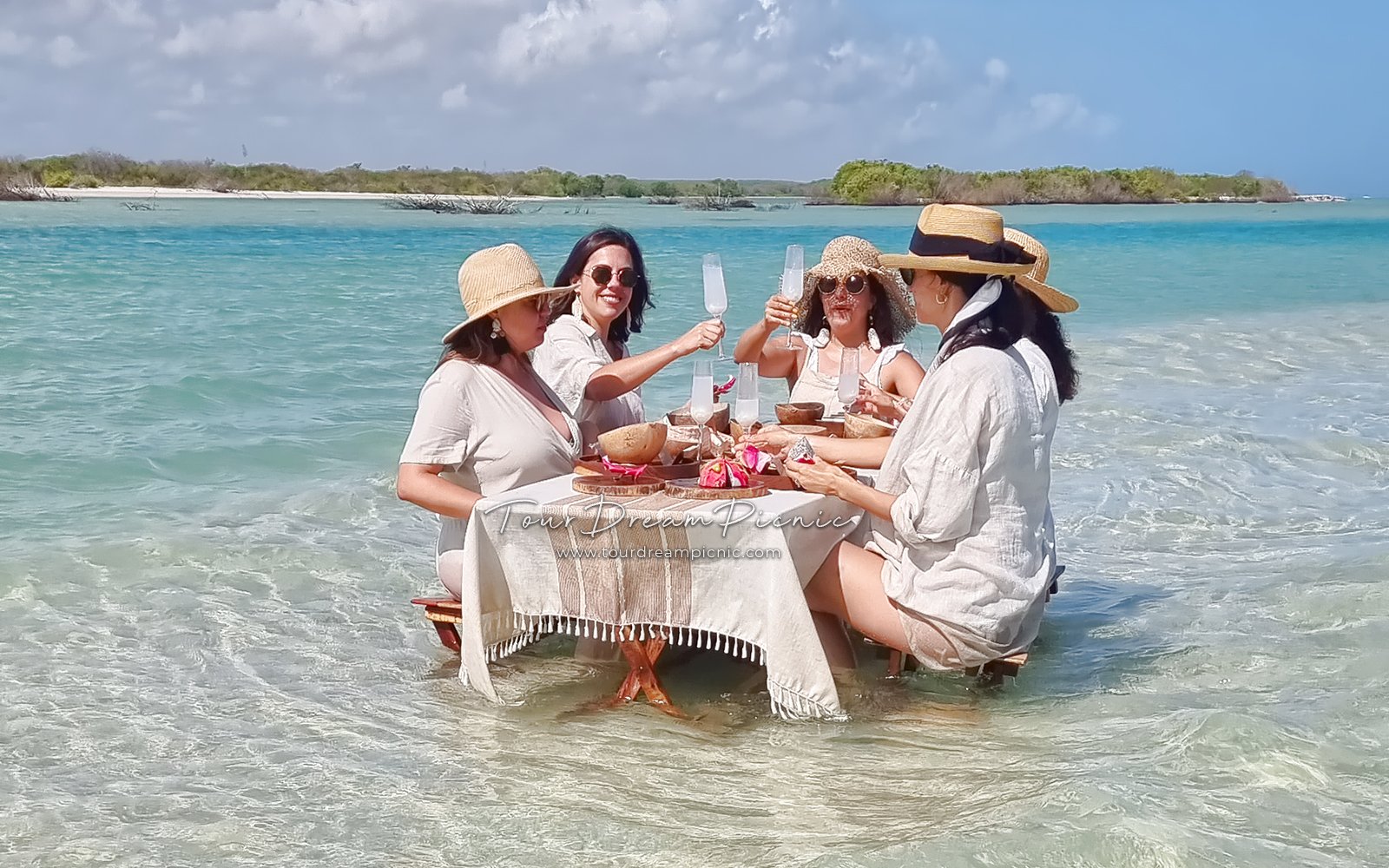 5 mujeres disfruten un picnic en una isla inundada o banco de arena hasta los tobillos en una playa virgen, vestidas con ropas de lino beige y están brindando con champaña. Picnic enn medio de mar, rodeado por agua, versión luxury en Sisal Yucatan Mexico